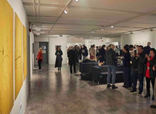 A large gallery space with yellow paintings on the left wall and groups of people gathered around tables and artworks throughout the room.