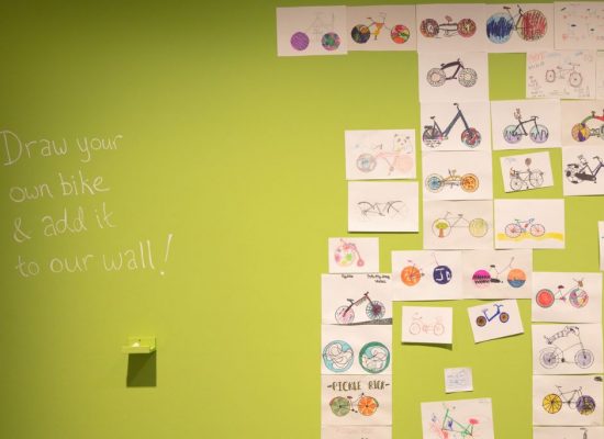 A green wall displays numerous hand-drawn bicycle illustrations arranged in a grid, with the text “Draw your own bike & add it to our wall!”.