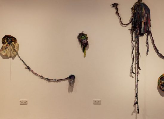 A wall installation features several colorful, textured fabric pieces connected by braided strands, arranged in an organic pattern.