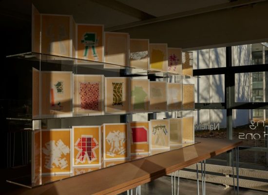 A display of colorful, layered paper artworks is arranged on clear shelves inside a gallery space, with sunlight streaming through large windows. The pieces feature abstract shapes and patterns in shades of yellow, orange, green, and red.