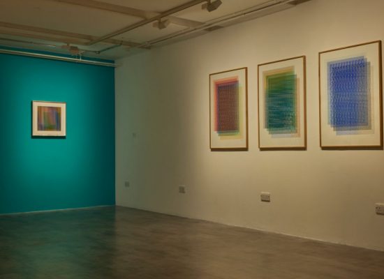 A gallery room features three framed abstract artworks with intricate patterns on one wall and a single striped artwork on a teal-colored wall.