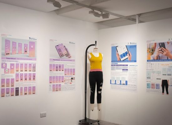 A gallery display features large posters on white walls showcasing app interface designs and informational graphics, primarily in purple and blue tones. In the center, a mannequin dressed in a yellow crop top and black leggings stands on a metal stand.