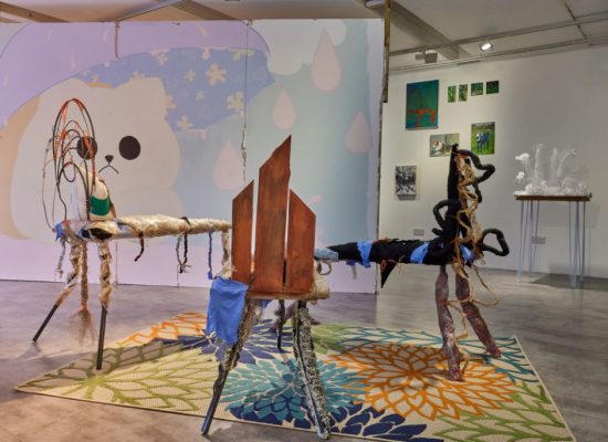A contemporary gallery installation features abstract sculptural pieces made of textured materials arranged on a colorful floral-patterned rug. The background includes large windows and a wall mural with soft pastel shapes and a cartoon-like face.