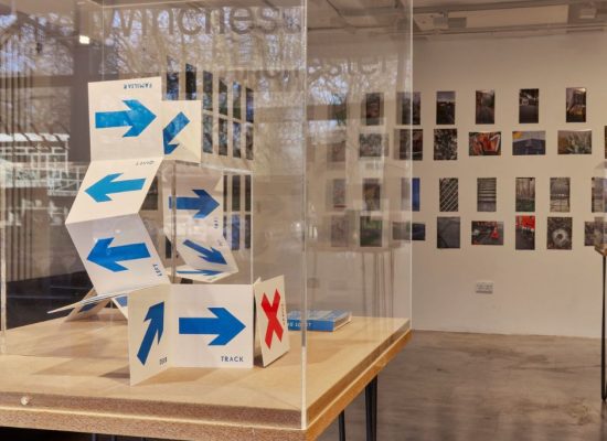 An art gallery display features a folded paper structure marked with blue arrows and a red 'X' inside a glass case. Behind it, a wall is covered with small photographs arranged in a grid, and another table with items is visible to the right.