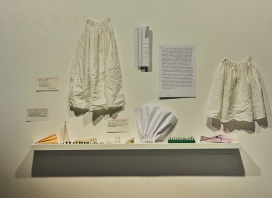 A white wall display features two gathered fabric garments, a sheet of text, and several small objects arranged on a narrow shelf, including folded paper pieces and miniature items. Labels with descriptions are mounted alongside the garments and objects.