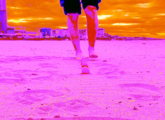 A person walks barefoot on a sandy beach with footprints visible in the foreground, set against a vivid, surreal color palette of pink sand and orange sky. Buildings and a cloudy horizon appear in the background.