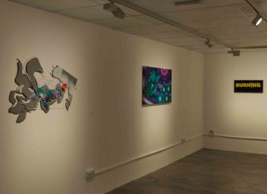 An art gallery with white walls displays three artworks: an abstract gray and colorful piece on the left, a vibrant purple and green painting in the center, and a black rectangular piece with the word “BURNING” in yellow on the right. The floor is gray, and the ceiling has track lighting illuminating the artworks.