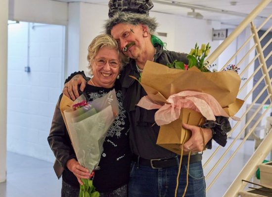 Two people stand indoors holding large bouquets of flowers wrapped in paper, near a staircase and a wall with artwork. The setting appears to be a gallery or event space.