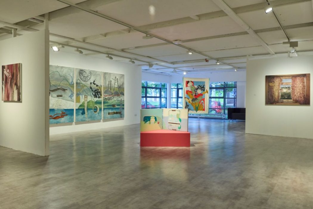 A bright gallery space with white walls displays large colorful paintings, including a multi-panel artwork and an abstract piece on a red pedestal in the center. Floor-to-ceiling windows in the background let in natural light.