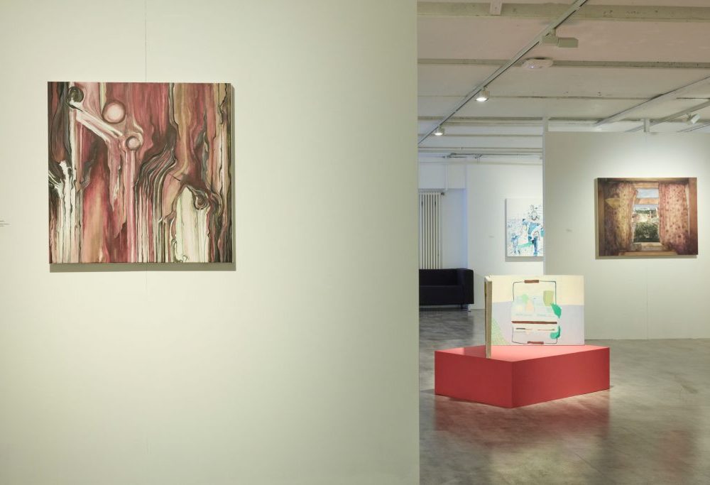 A close-up view of a gallery wall featuring an abstract painting in shades of red and brown. In the background, a red pedestal holds a small artwork, and other paintings are visible on adjacent walls.