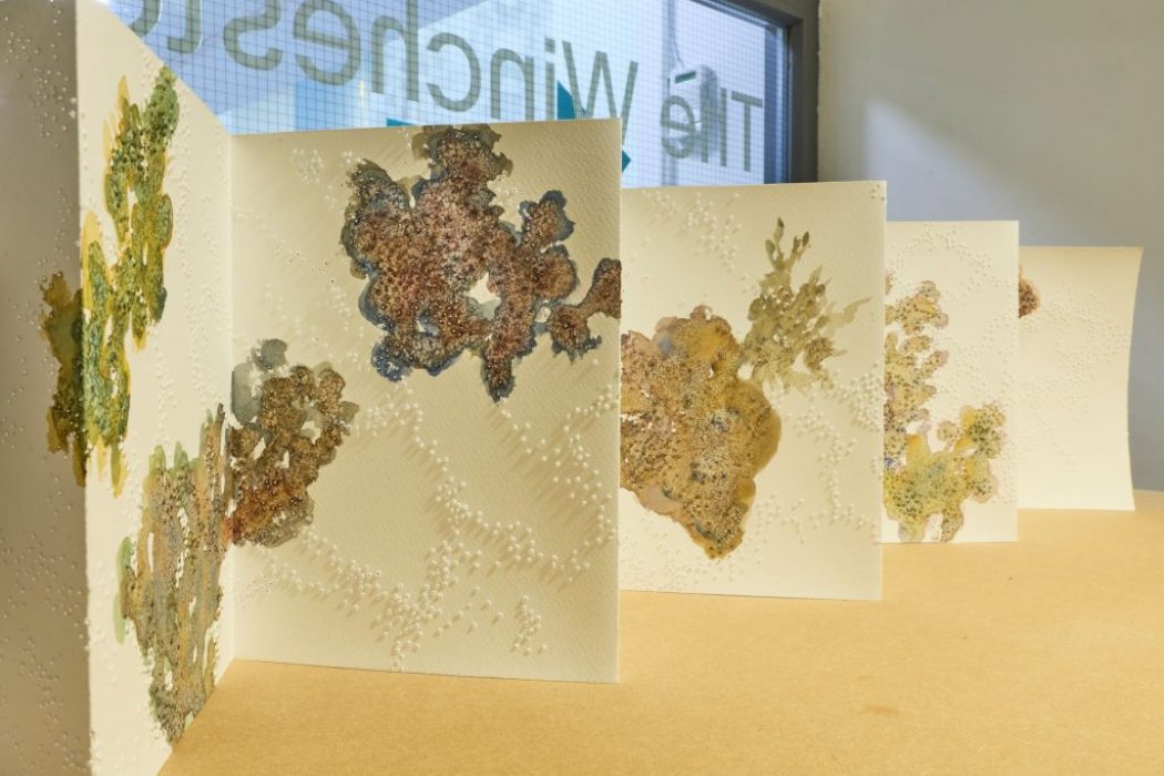 A folded paper artwork with textured surfaces and organic patterns in earthy tones is displayed on a wooden table. The background shows a glass door with partial text reading “The Winchester.”