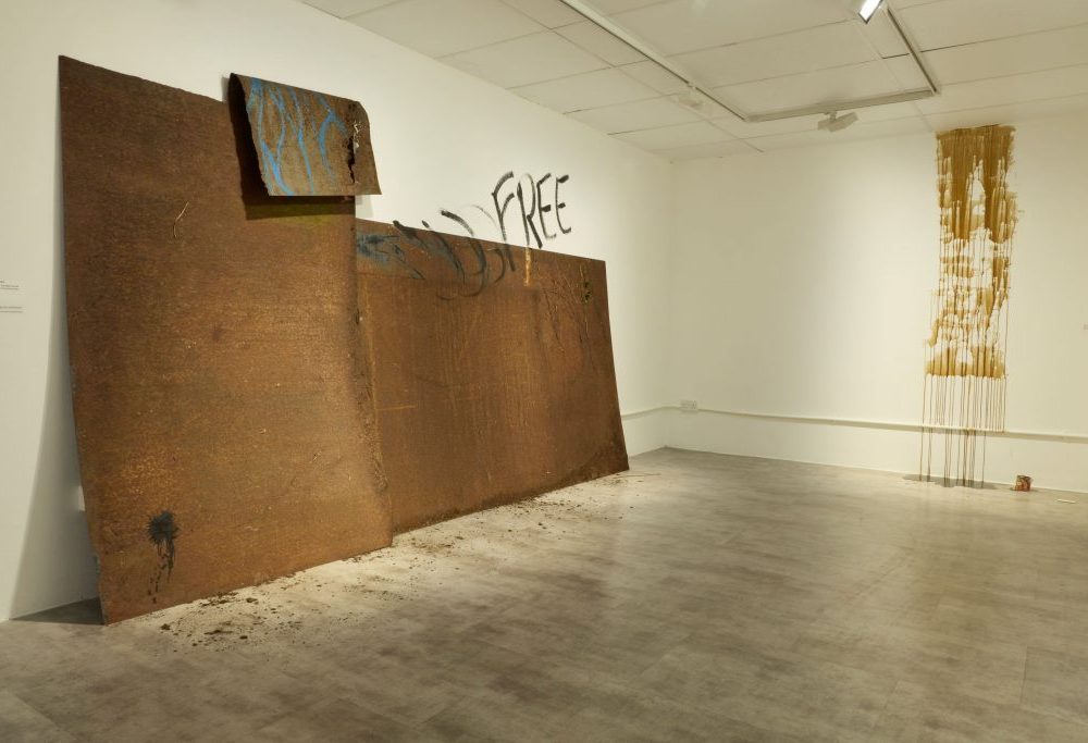 A gallery corner with a large rust-colored metal installation leaning against the wall, partially painted with blue and black marks and the word “FREE.” A vertical artwork with dripping gold paint hangs on the adjacent wall.