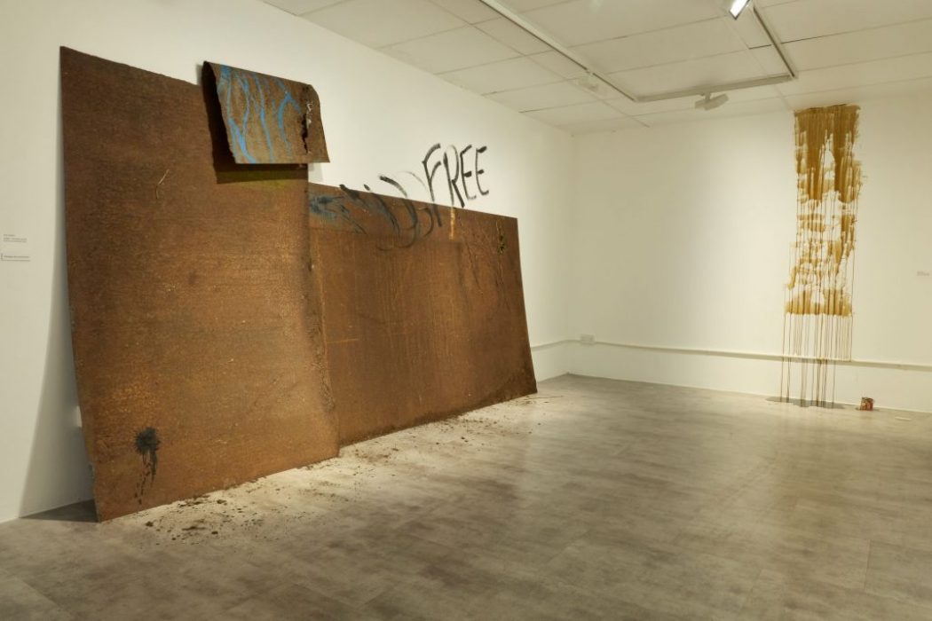 A gallery corner with a large rust-colored metal installation leaning against the wall, partially painted with blue and black marks and the word “FREE.” A vertical artwork with dripping gold paint hangs on the adjacent wall.