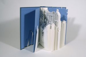 An altered copy of Wuthering Heights stands partially open, its pages sculpted and cut away into a dense, textured landscape, with a blue cover framing the carved interior.