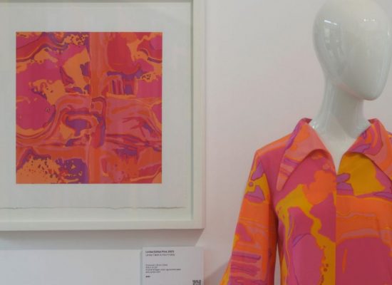A framed textile print in vivid pink and orange abstract patterns hangs beside a mannequin wearing a matching patterned garment. The display highlights the connection between the printed artwork and the finished clothing design.