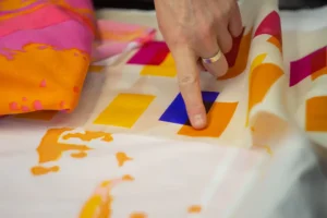 A hand points to a square of bright colour on a piece of patterned fabric laid out on a table. The fabric includes vivid blocks of yellow, orange, pink, and blue alongside flowing painted designs.