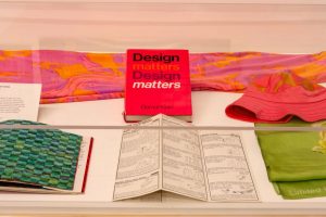 A display case contains a bright pink book titled Design Matters alongside woven textiles, sewing instructions, and colourful patterned fabrics. The objects are arranged neatly with a pink and orange fabric piece placed behind the book.