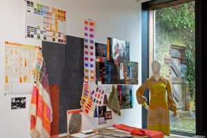 A studio display shows textile samples, colour charts, sketches, and fabric swatches pinned to a wall above a wooden table. A mannequin wearing a brightly patterned garment stands beside the workspace next to a large window.