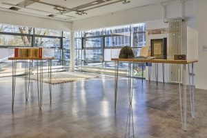 A bright gallery space with large windows displays two tall tables holding books and archival objects in transparent cases. Sunlight casts reflections across the floor and surrounding exhibits.