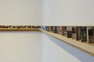 A narrow wooden shelf runs along the corner of a white wall, holding a sequence of small photographic prints. The photos form a continuous horizontal strip showing outdoor architectural scenes.