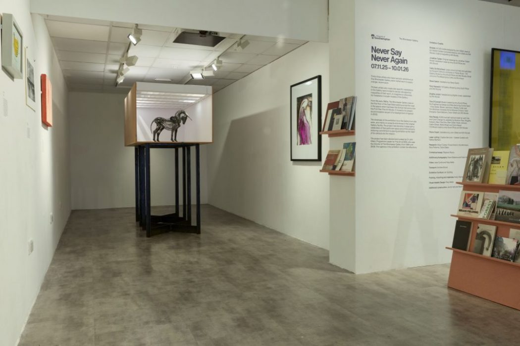 A gallery corridor features a large box-like display structure containing a skeletal animal sculpture elevated on tall blue legs. Nearby walls hold framed artworks, bookshelves, and exhibition text titled “Never Say Never Again.”