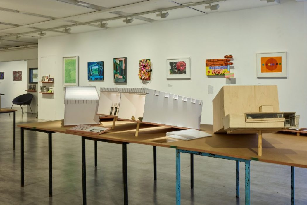 A row of architectural models is displayed on long tables in a brightly lit gallery, with vibrant paintings and prints mounted on the wall behind them. The models vary in shape and material, creating a visually layered exhibition.