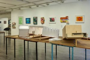 A row of architectural models is displayed on long tables in a brightly lit gallery, with vibrant paintings and prints mounted on the wall behind them. The models vary in shape and material, creating a visually layered exhibition.