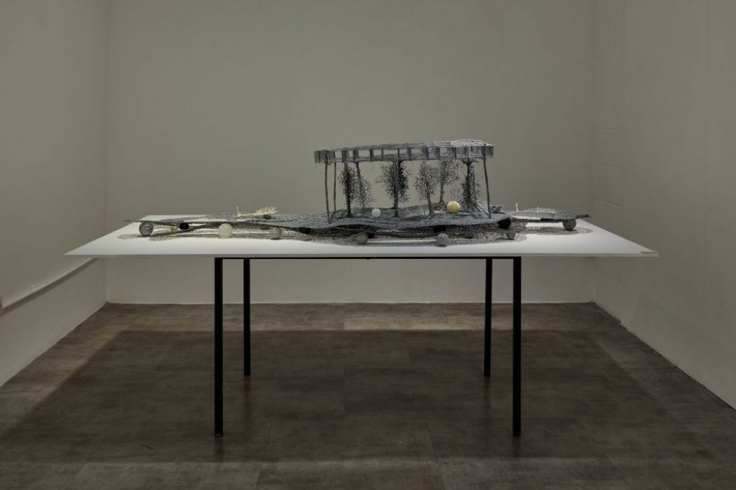 A sculptural installation made of delicate, metallic-looking components is arranged across a white table in a minimalist room. The artwork resembles an intricate landscape with raised structures and fine details.