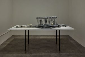 A sculptural installation made of delicate, metallic-looking components is arranged across a white table in a minimalist room. The artwork resembles an intricate landscape with raised structures and fine details.