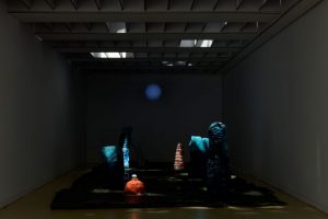 A dimly lit installation features tall blue and orange sculptural forms arranged on a dark textured surface inside a narrow room. A soft blue circular light is projected onto the back wall, creating an atmospheric scene.