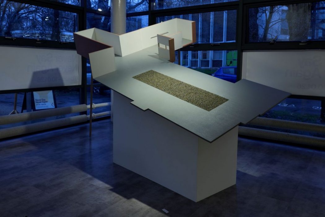 A large architectural model with angled surfaces is displayed on a pedestal in front of floor‑to‑ceiling windows. A rectangular textured section is embedded in the centre of the tilted plane, illuminated by cool indoor lighting.