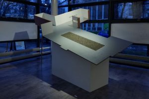 A large architectural model with angled surfaces is displayed on a pedestal in front of floor‑to‑ceiling windows. A rectangular textured section is embedded in the centre of the tilted plane, illuminated by cool indoor lighting.