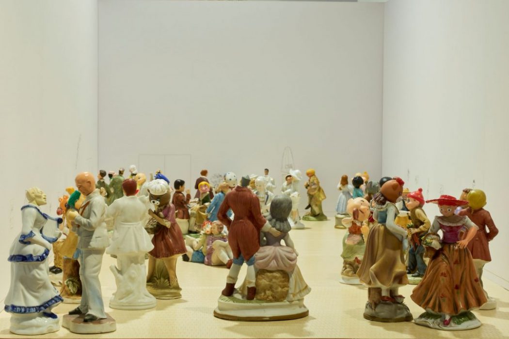 A large group of small ceramic figurines is arranged across a pale floor, each posed as if interacting or dancing. The figures fill the space from foreground to background within white gallery-style walls.