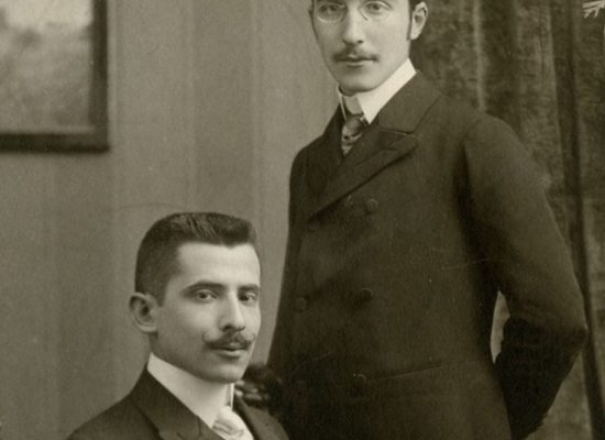 A sepia-toned vintage studio portrait shows one person seated in an ornate wooden chair while another stands beside them, both dressed in formal early 20th‑century attire. The background features a draped curtain, a framed wall picture, and a potted plant.