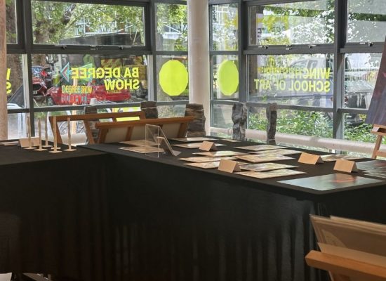 An art gallery corner with black-covered tables displaying prints and small sculptures, and a large figurative painting on an easel near a window.