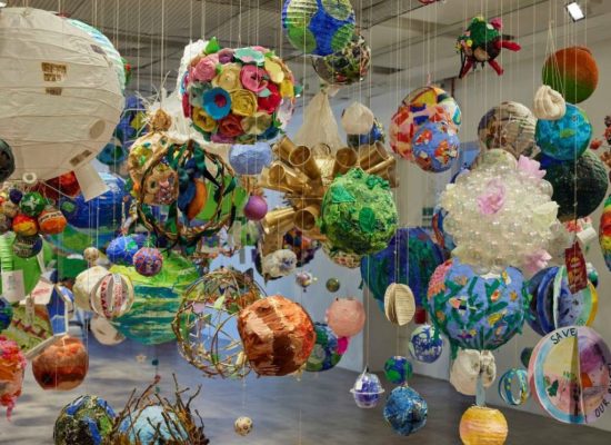 An art installation features numerous colorful spherical objects suspended from the ceiling by thin strings. The spheres vary in size and design, creating a vibrant floating display in a spacious gallery.