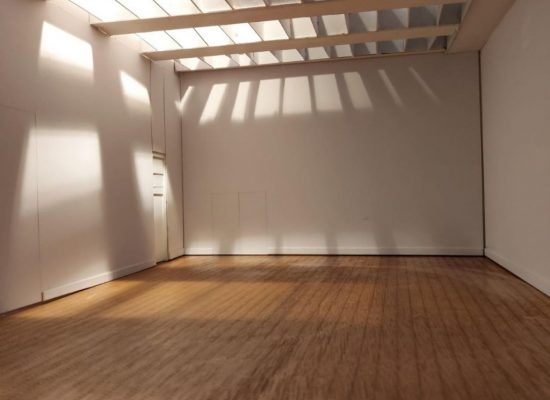 **Alt text:** An empty white gallery space with a wooden floor is lit by sunlight streaming through a slatted ceiling, casting linear shadows across the walls and floor. A closed door is visible on the left, emphasising the quiet, minimal interior.