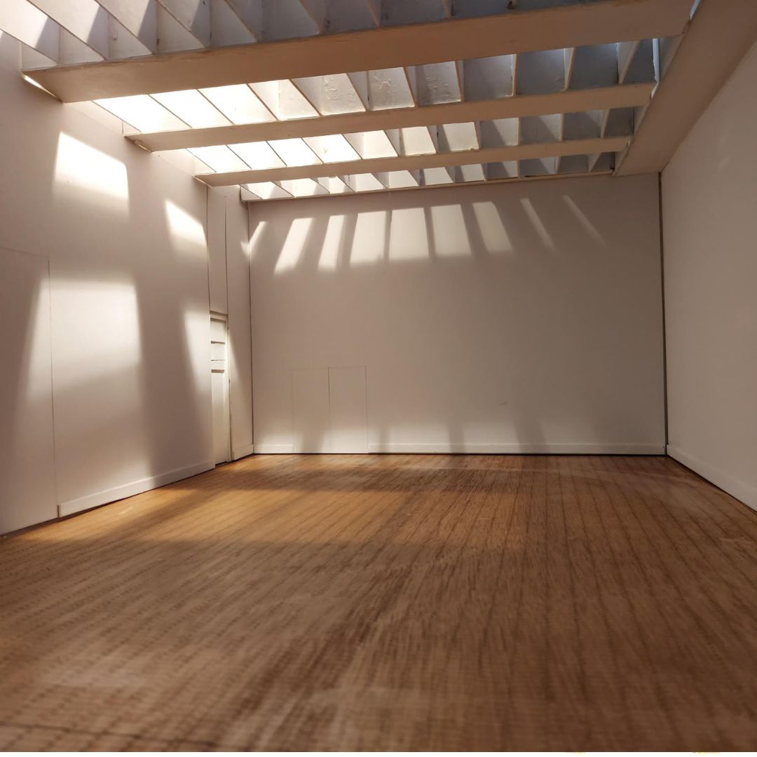 **Alt text:** An empty white gallery space with a wooden floor is lit by sunlight streaming through a slatted ceiling, casting linear shadows across the walls and floor. A closed door is visible on the left, emphasising the quiet, minimal interior.