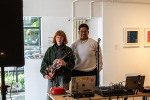 Two performers stand behind a table of audio equipment, one holding a violin and the other standing near a microphone. Framed artworks hang on the wall behind them in a bright gallery interior.