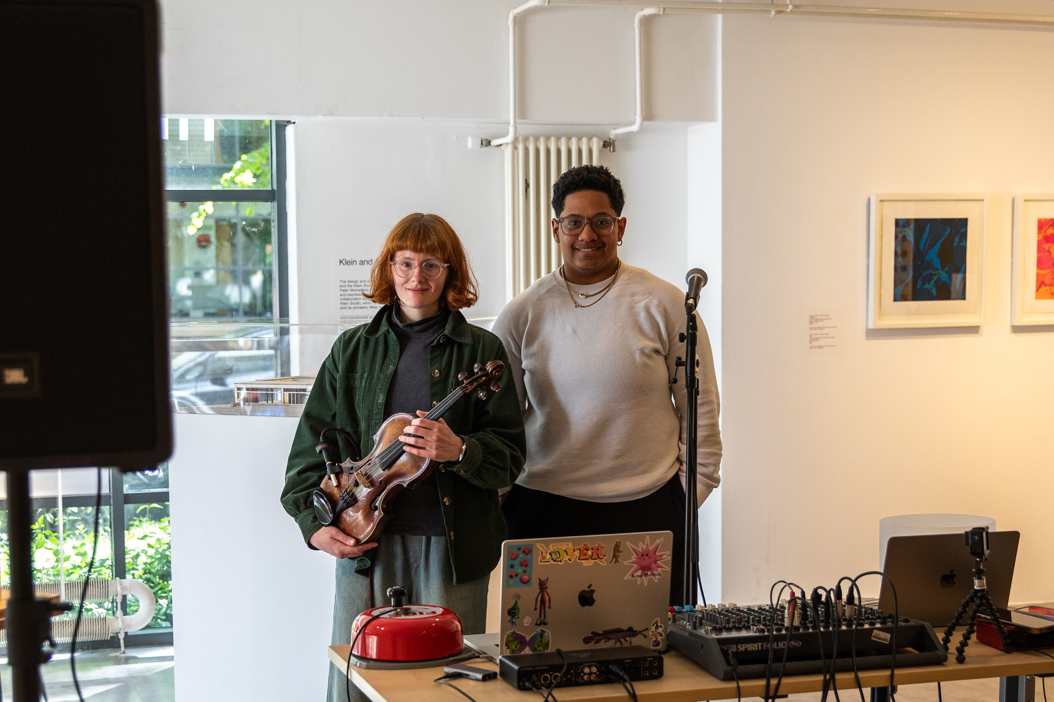 Two performers stand behind a table of audio equipment, one holding a violin and the other standing near a microphone. Framed artworks hang on the wall behind them in a bright gallery interior.