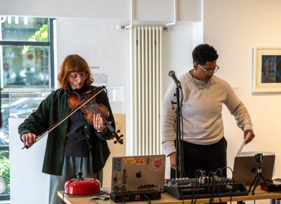 Two people perform live music indoors, with one playing a violin and the other adjusting audio equipment beside a microphone stand. A table holds a laptop, mixer, cables and small instruments, set within a gallery space.