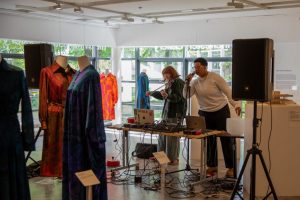A wider view of the performance shows two musicians behind a table of sound equipment, flanked by speakers and surrounded by displayed garments on mannequins. Large windows and gallery labels indicate an exhibition setting.