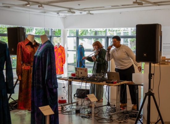 A wider view of the performance shows two musicians behind a table of sound equipment, flanked by speakers and surrounded by displayed garments on mannequins. Large windows and gallery labels indicate an exhibition setting.