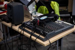 A table holds a mixing desk, speakers, headphones and a vinyl record, with multiple cables running across the surface. A laptop and water bottle sit nearby in a busy technical workspace.