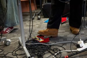 A close‑up shows a foot pressing a red effects pedal on the floor, surrounded by tangled audio cables and power leads. Table legs, wheels and nearby shoes indicate a live performance setup.