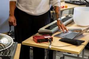 Hands rest on a laptop trackpad beside a small keyboard, audio interface and a translucent bowl on a table. Cables and electronic equipment suggest live sound or music production.