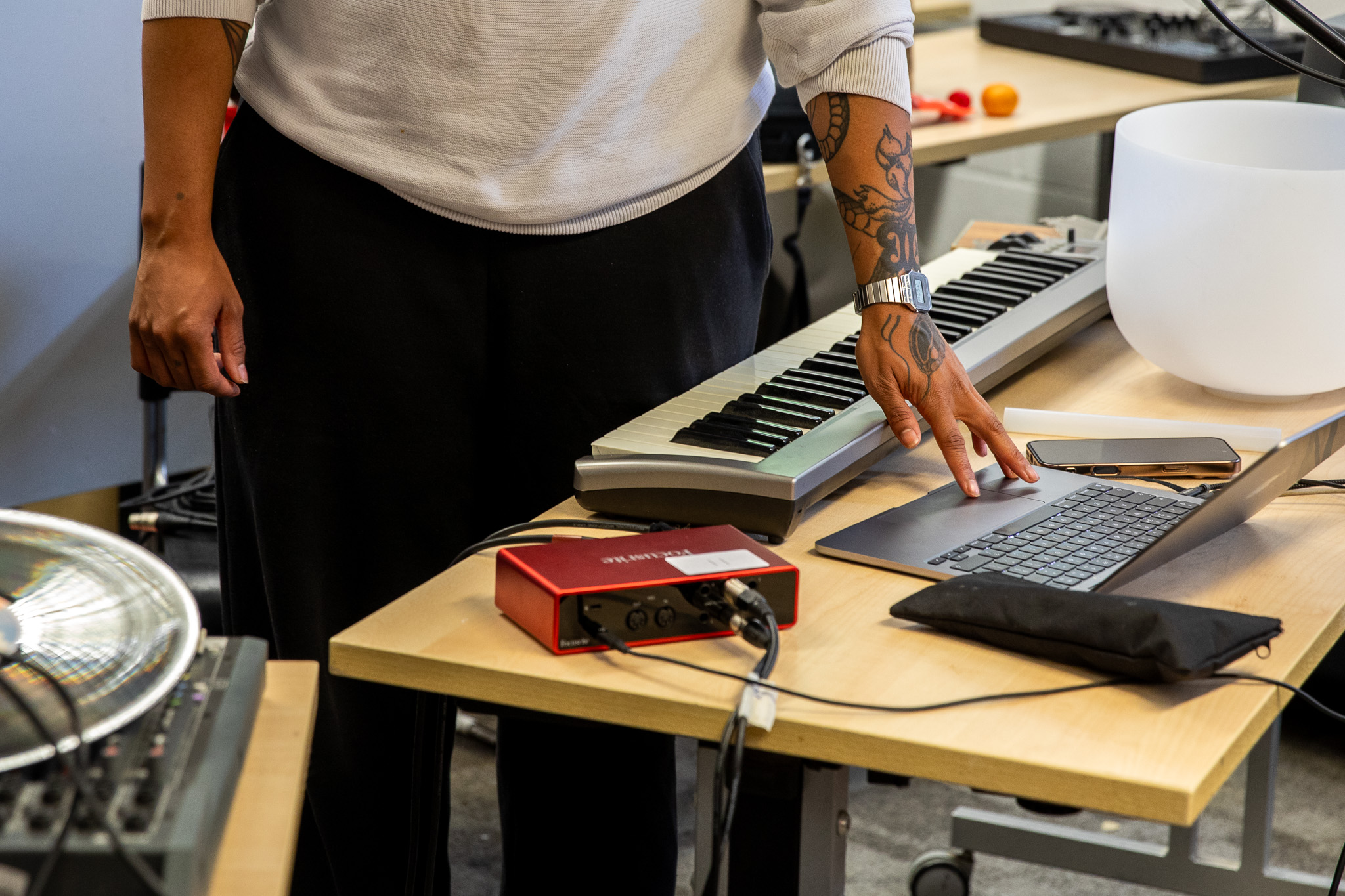 Hands rest on a laptop trackpad beside a small keyboard, audio interface and a translucent bowl on a table. Cables and electronic equipment suggest live sound or music production.