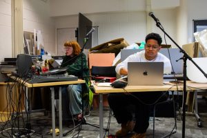 Two people sit at separate tables using laptops and audio equipment in a studio space filled with cables, microphones and speakers. Stacked boards and materials in the background indicate a workshop or preparation area.