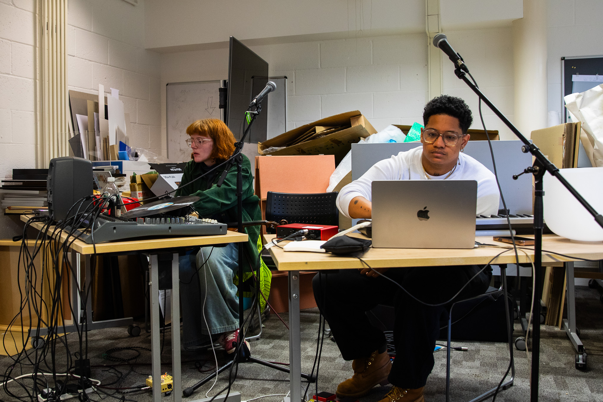 Two people sit at separate tables using laptops and audio equipment in a studio space filled with cables, microphones and speakers. Stacked boards and materials in the background indicate a workshop or preparation area.