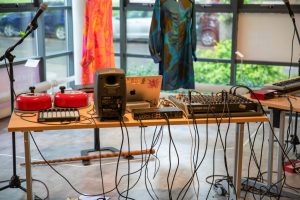 A table is set up with audio equipment including speakers, a mixer, a laptop, cables, and two red hand bells. Colourful garments hang in the background in a bright gallery space with large windows.
